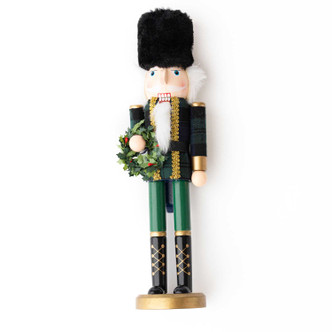 Hand-painted wood nutcracker in a black fur hat and dark green plaid-inspired uniform, holding a holiday wreath