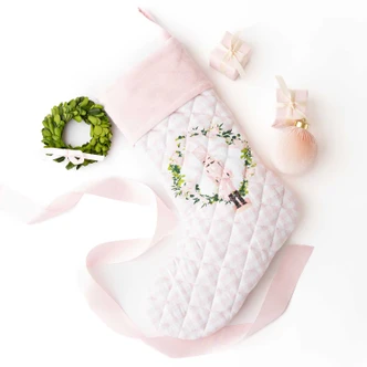 Blush pink quilted Christmas stocking with a velvet cuff, wreath, and nutcracker embroidery, hung on a mantel (alternate angle)
