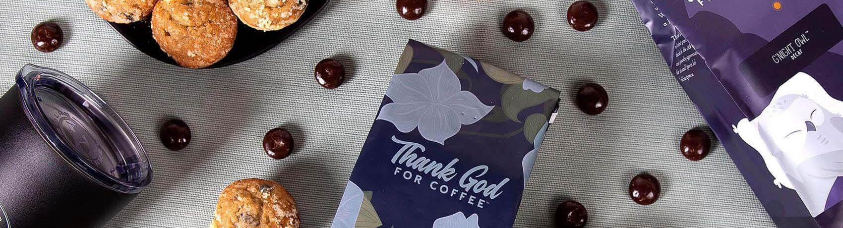 Image of Thank God For Coffee dark roast bags on a table with cookies and chocolates.