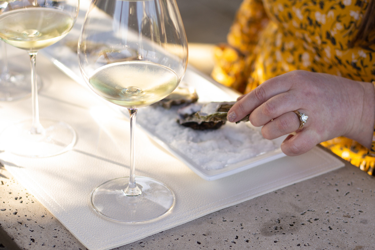 table with two glasses of white wine and a hand holding an oyster on a plate full of salt