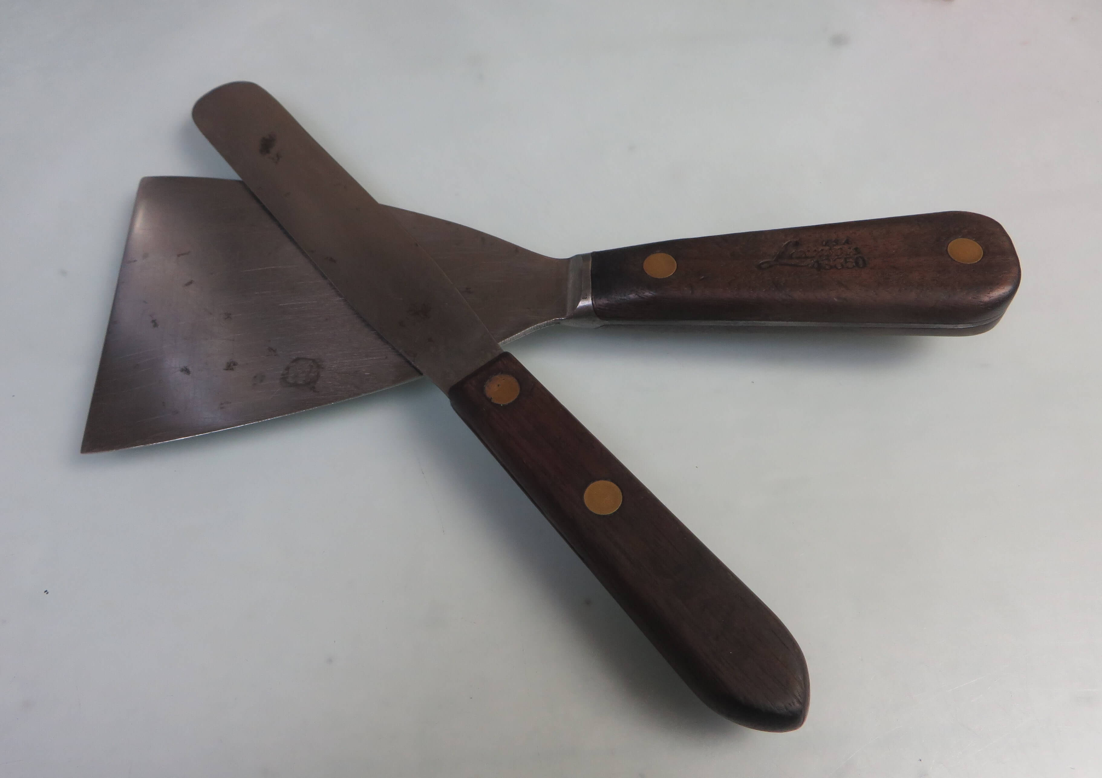 Two ink knives with wooden handles and metal blades crossed on a white work surface. The blades show signs of wear and ink residue, indicating frequent use in printmaking or ink mixing.
