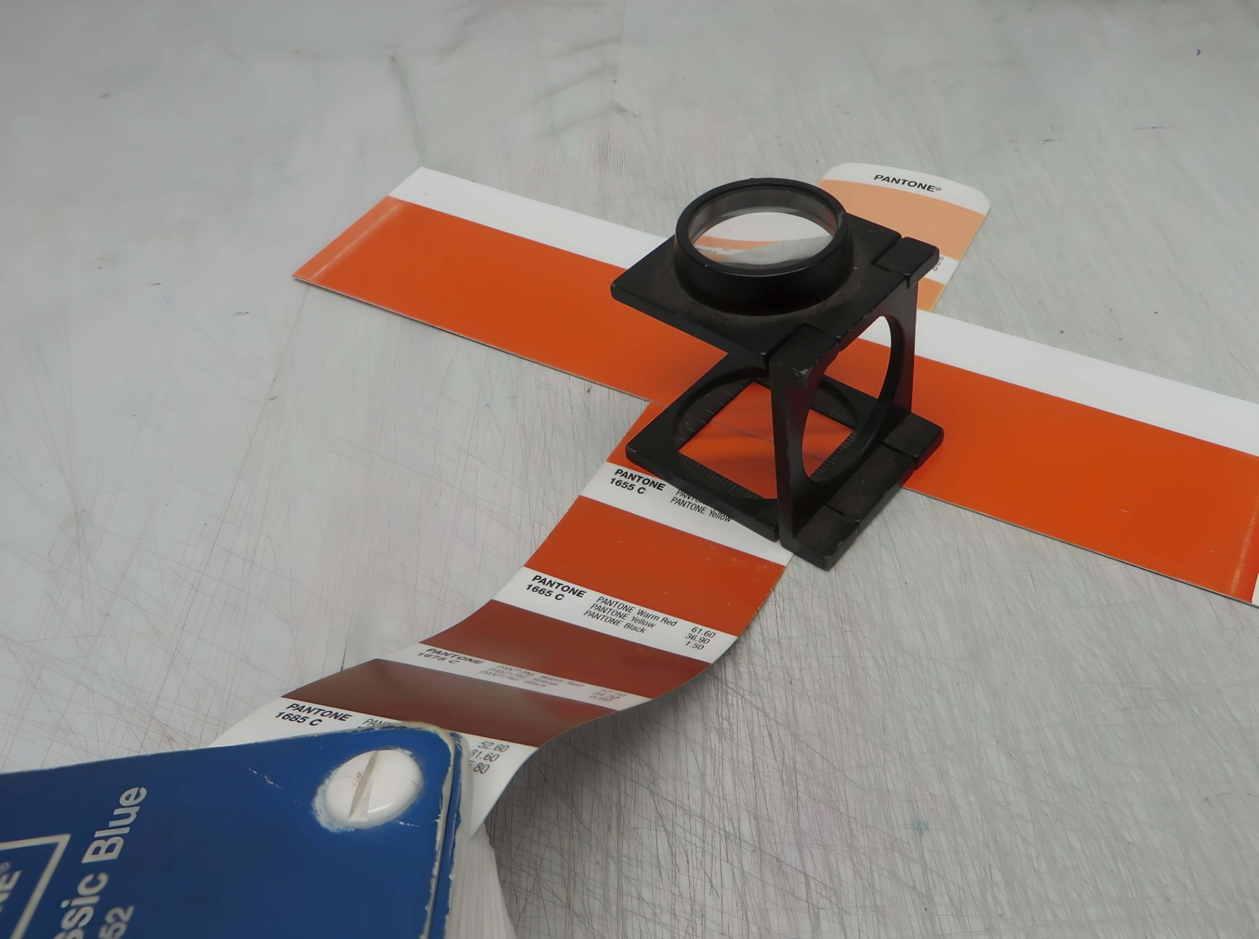 A Pantone color swatch book displaying shades of orange and red, placed on a work surface. A black folding magnifier (loupe) is positioned over a printed orange color sample for close inspection.