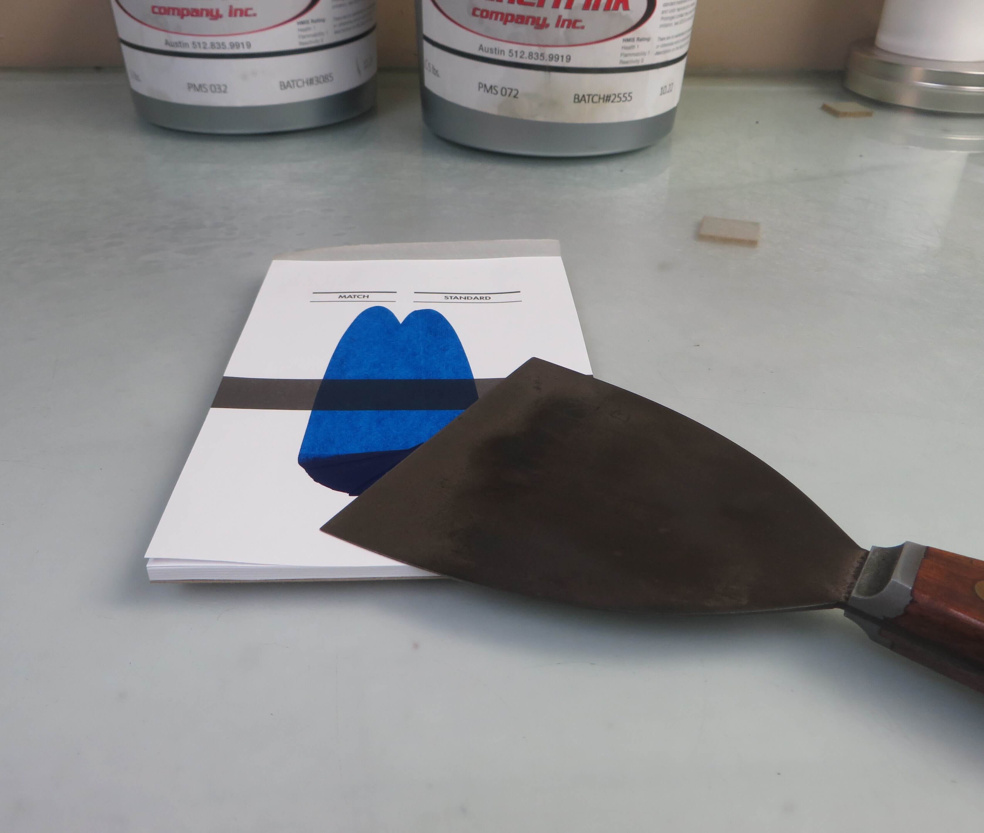A putty knife with a wooden handle resting on a printed color match sheet with a blue ink swatch. In the background, two cans of printing ink are visible on a work surface.
