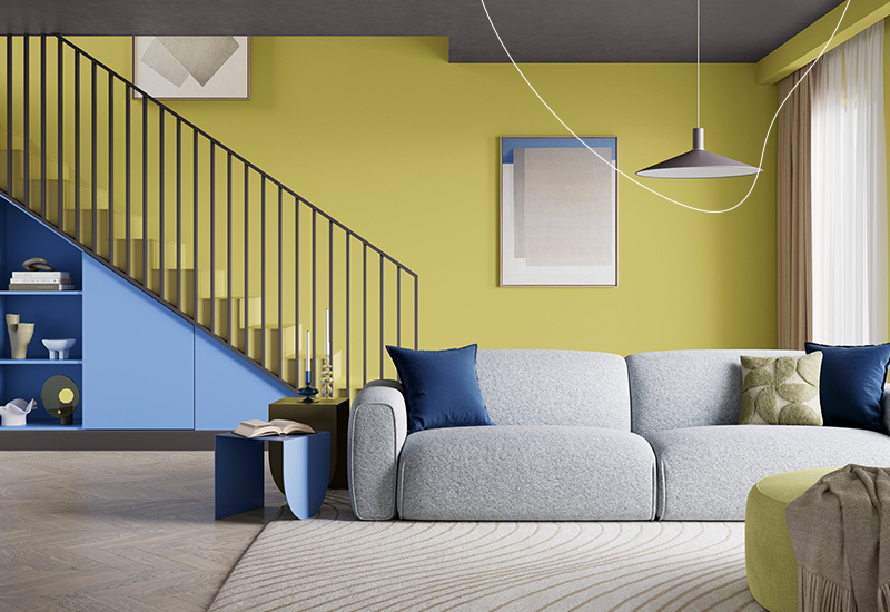 Moderne woonkamer met geelgroene muur in de kleur ‘Secret Safari’, grijze bank, blauwe trapkast onder de trap en veel daglicht door grote gordijnen.