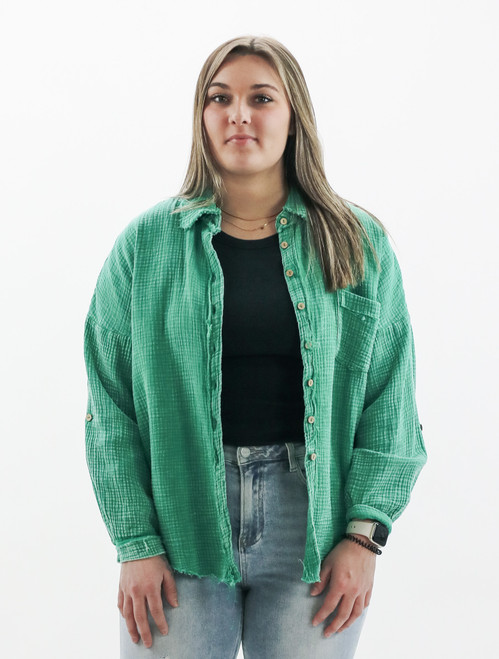 Discover Trendy Threads Boutique's spring collection with the new arrival: a vibrant green oversized gauze top. This top exudes a fresh, casual vibe with its rich textured fabric, featuring frayed seams and hem for an edgy twist. Styled over a black tee and paired with light-wash jeans, the top boasts a full button front closure and a practical chest pocket. The garment's relaxed fit is ideal for a comfortable yet stylish seasonal transition, modeled by a woman accessorized with a smartwatch, showcasing the top's versatility and laid-back charm.