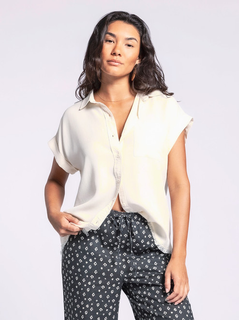 Confident woman modeling the Ambrose Top in an ivory shade, featuring a full button front, fold-over collar, and cuffed short sleeves, paired with patterned drawstring pants. The top's frayed hem adds a casual flair.