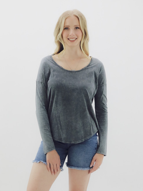 A woman wearing a soft cotton long sleeve tee from Mustard Seed in a gray color, featuring a wide rounded neckline, drop shoulders, and curved hem, paired with denim shorts.