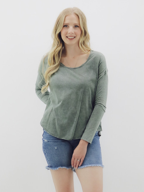 A woman wearing a soft cotton long sleeve tee from Mustard Seed in a green color, featuring a wide rounded neckline, drop shoulders, and curved hem, paired with denim shorts.