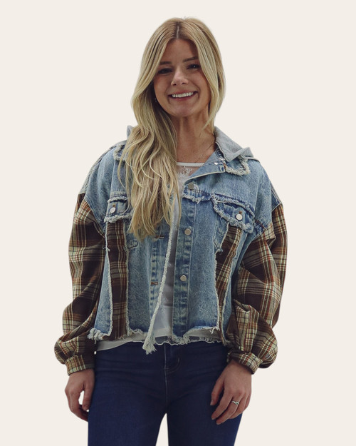Front view of a woman wearing a cropped denim jacket with brown plaid sleeves, raw hem, distressed details, and a gray hood over a white top.