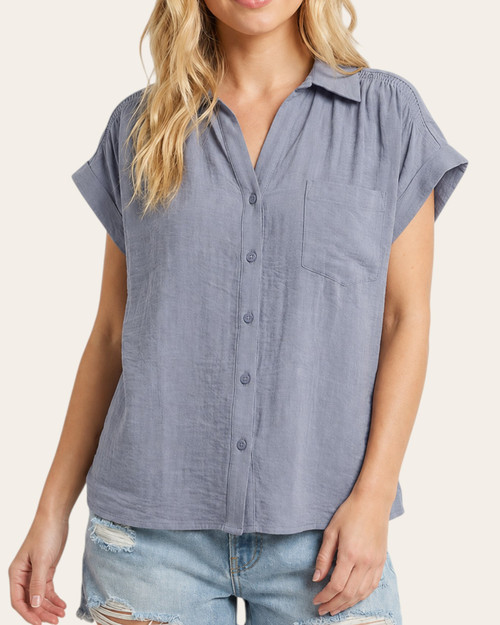 Woman facing forward wearing a denim blue smocked shoulder short-sleeve blouse with button front details.