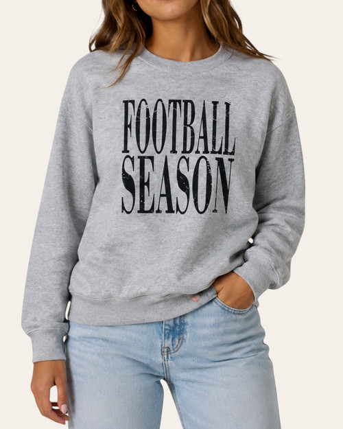 Woman wearing a cozy grey graphic sweatshirt with the words Football Season printed on the front, styled with light wash jeans.