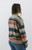 Right back side view of a woman in a relaxed-fit, multicolored striped sweater from Trendy Threads Boutique's Spring collection. The sweater features a combination of soft pastel and earth tones in horizontal stripes, with a V-neckline and ribbed cuffs and hem. She is wearing light blue denim jeans and has straight, shoulder-length hair. The background is a simple, clean white, emphasizing the sweater's colors.