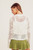 View from the back of a woman wearing a white open-knit cardigan with a floral daisy design, featuring a buttoned front and ribbed hem, matched with a playful olive green flared mini skirt.

