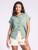 Front view of a woman smiling subtly, wearing the Ambrose Top by Thread & Supply in light green, with a front button closure, classic collar, cuffed short sleeves, and a frayed hemline. She's also wearing white shorts, and the top is casually tucked in at the front.