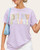 Front view of a model wearing a soft lilac graphic tee with the word PRAY in pastel floral and gingham patchwork letters, available at our Roscoe SD boutique.