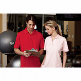 A man in a red polo and a woman in a pink polo stand together, holding a clipboard in a gym setting.