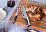 A carving set with a knife and fork on a wooden cutting board, featuring cooked meat. The utensils have black handles.