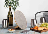 A round silver and wood Bluetooth speaker with a textured surface, accompanied by a wine bottle, glasses, and snacks.