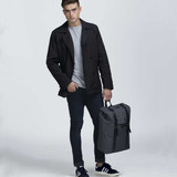 A grey front-side backpack held by a person wearing a black coat, grey shirt, and dark jeans. A grey front-side backpack held by a person wearing a black coat, grey shirt, and dark jeans.