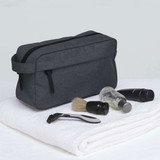 A dark grey toiletry bag with grooming items including a razor, shaving brush, and travel-sized bottles, placed on a white towel.