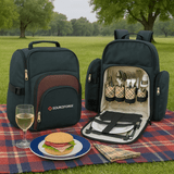 A dark green picnic backpack displaying cutlery, plates, and a sandwich on a blue plate, set on a picnic blanket.