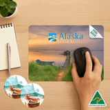 A mouse mat featuring a scenic landscape with a logo, paired with two circular coasters, on a wooden desk.