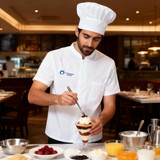 A chef wearing a white short-sleeve single-breasted jacket and hat prepares a dessert with various ingredients on the table.