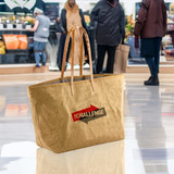 A large, tan kraft paper tote bag with handles and a logo displayed in a shopping area. A large, tan kraft paper tote bag with handles and a logo displayed in a shopping area.