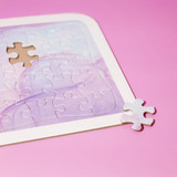 A jigsaw puzzle with pastel purple shades and one piece detached on a pink background. It features rounded corners and a logo.