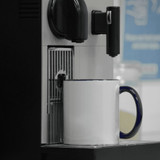 A two-tone ceramic mug with a white body and blue handle sits under a coffee machine. It has a logo on the side.