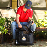 A soft roll-top cooler bag in black with a logo, being accessed by a person in a red shirt and cap.