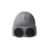 A grey beanie featuring large black goggles on the front, with a textured knit design. It includes a logo.