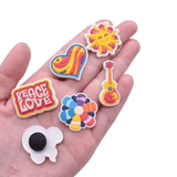 A hand holding six colourful PVC shoe charms featuring designs like a heart, sun, guitar, and flowers.