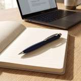 A blue ballpoint pen lies on an open notebook, with a laptop in the background. The setting is well-lit and modern.