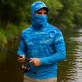 A blue camo fishing hoodie with a hood and face covering, featuring a logo, worn by a person near a river.