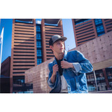 A young man wearing a grey and black trucker cap, styled with a denim jacket, standing in an urban environment.