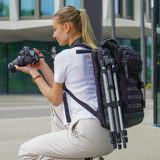 A woman with a black Swissdigital Berg XL Backpack crouches, holding a camera. The backpack has a tripod attached.