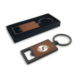 A drink bottle opener with a square wooden panel and a metallic keyring, presented in a black box.
