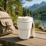 A reusable double wall coffee cup in white, placed on a wooden table near a natural landscape, with a bag nearby.