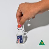 A hand holds a clear tear drop acrylic keychain featuring the Australian flag. The keychain has a silver ring and a logo.