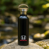 A drink bottle in matte black with a metal lid and a logo on the front, set against a natural background.