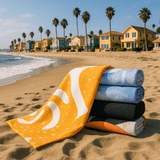 A stack of five bath towels in various colours, including yellow, blue, and black, on a sandy beach with palm trees and houses in the background.