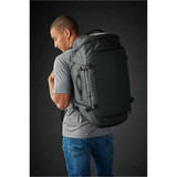 A man wears a large black duffle bag on his back, paired with a grey t-shirt and blue jeans. The bag features straps and a logo.