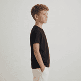 A child stands in profile wearing a black essential tee and white shorts, with hands in pockets.