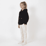 A young girl stands wearing a black hooded sweatshirt and white pants, with a light background. The hoodie has a logo.
