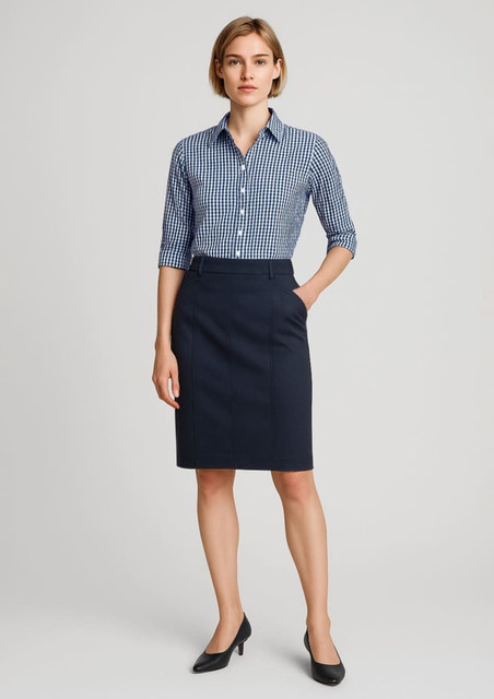 A woman wearing a navy mid waist chino skirt and a blue checked shirt, standing with hands in pockets.