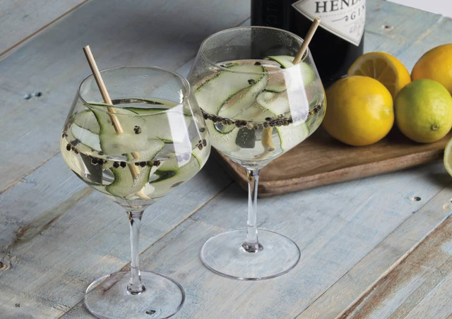 Two elegant glass goblets filled with a clear drink, garnished with cucumber slices and peppercorns, on a rustic wooden surface.