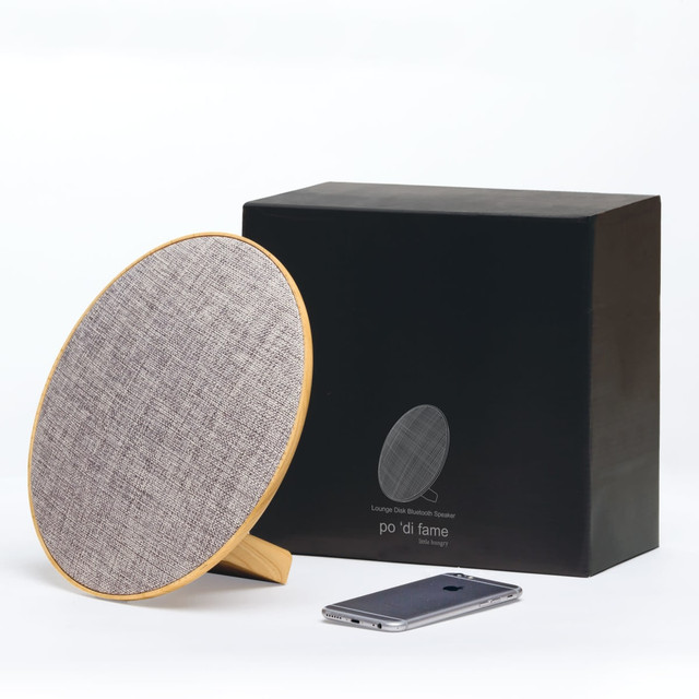 A round grey Bluetooth speaker with a wooden base next to a smartphone, accompanied by a black box packaging.