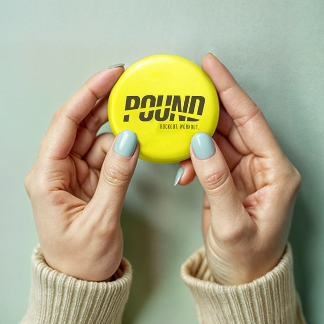 A round 75mm button badge in bright yellow, held between two hands, featuring a logo.