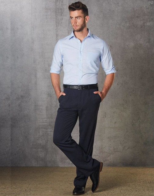 A man in a light blue shirt and black permanent press pants stands with hands in pockets against a grey backdrop.